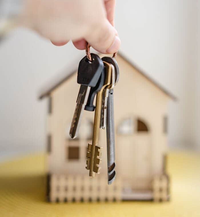 Close-up of a hand holding keys with a miniature wooden house in the background, symbolizing real estate investment.